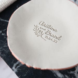 May include: A white ceramic dish with a rough, pink rim. The dish has the names "Christina and Brand" and the date "11.04.22" engraved, along with a small floral design. The dish is on a black and white marble surface.
