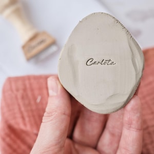 May include: A light gray, irregularly shaped ceramic piece with the name "Carlota" inscribed in cursive. The piece is held in a hand, with a wooden tool and a pink cloth visible in the background. The ceramic has a textured surface.