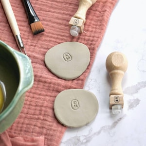 May include: A craft scene featuring pottery tools and materials. Two clay stamps with wooden handles and clear rollers are next to two clay pieces with stamped initials. A paintbrush and other tools are on a pink cloth.