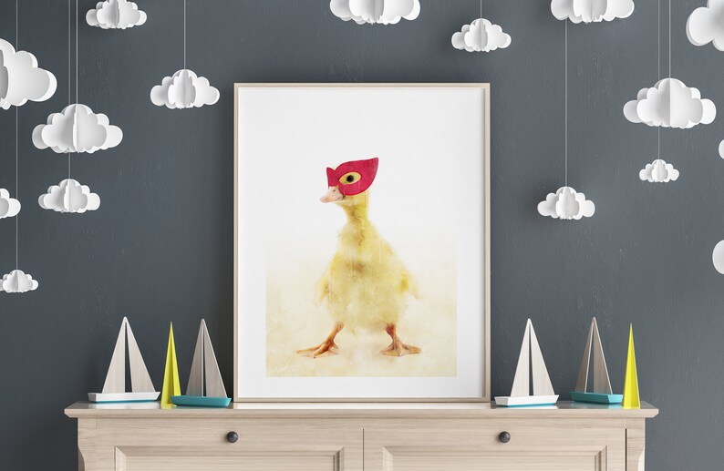 May include: A yellow duckling wearing a red superhero mask stands on a white background. The duckling is looking to the left of the image. The image is framed and hanging on a wall with white paper clouds hanging from the ceiling.