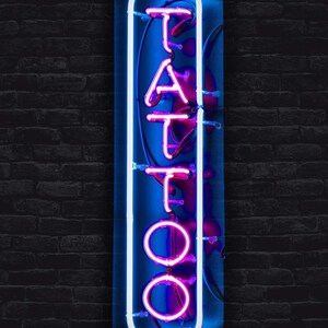May include: A neon sign with the word "TATTOO" in pink neon letters on a blue background. The sign is framed in white neon and is mounted on a brick wall.