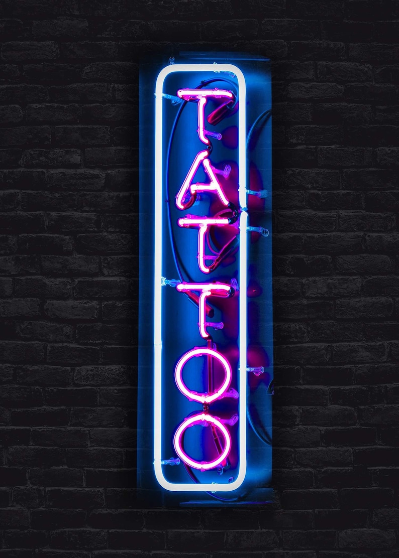 May include: A neon sign with the word "TATTOO" in pink neon letters on a blue background. The sign is framed in white neon and is mounted on a brick wall.