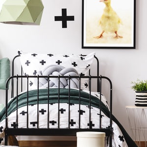 May include: A black metal bed frame with a white and black patterned duvet cover and a green throw blanket. A grey knotted pillow sits on the bed. A framed print of a duck wearing a red mask hangs on the wall above the bed.