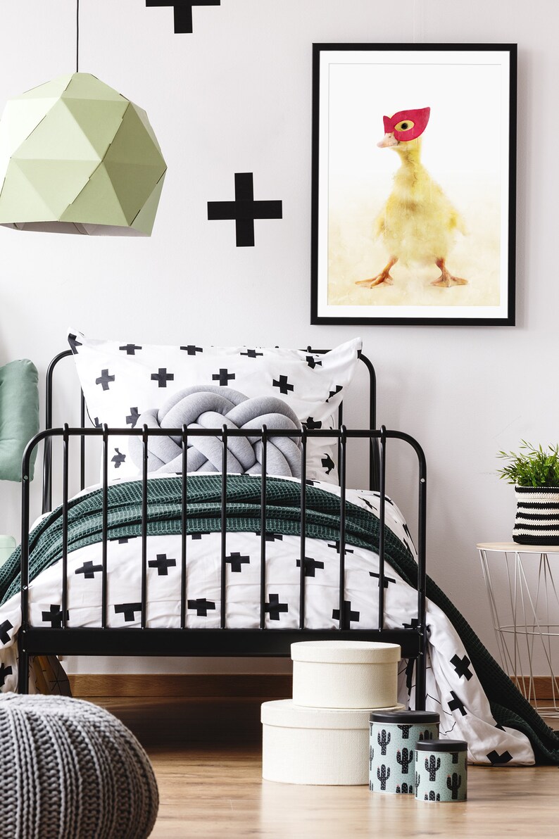 May include: A black metal bed frame with a white and black patterned duvet cover and a green throw blanket. A grey knotted pillow sits on the bed. A framed print of a duck wearing a red mask hangs on the wall above the bed.
