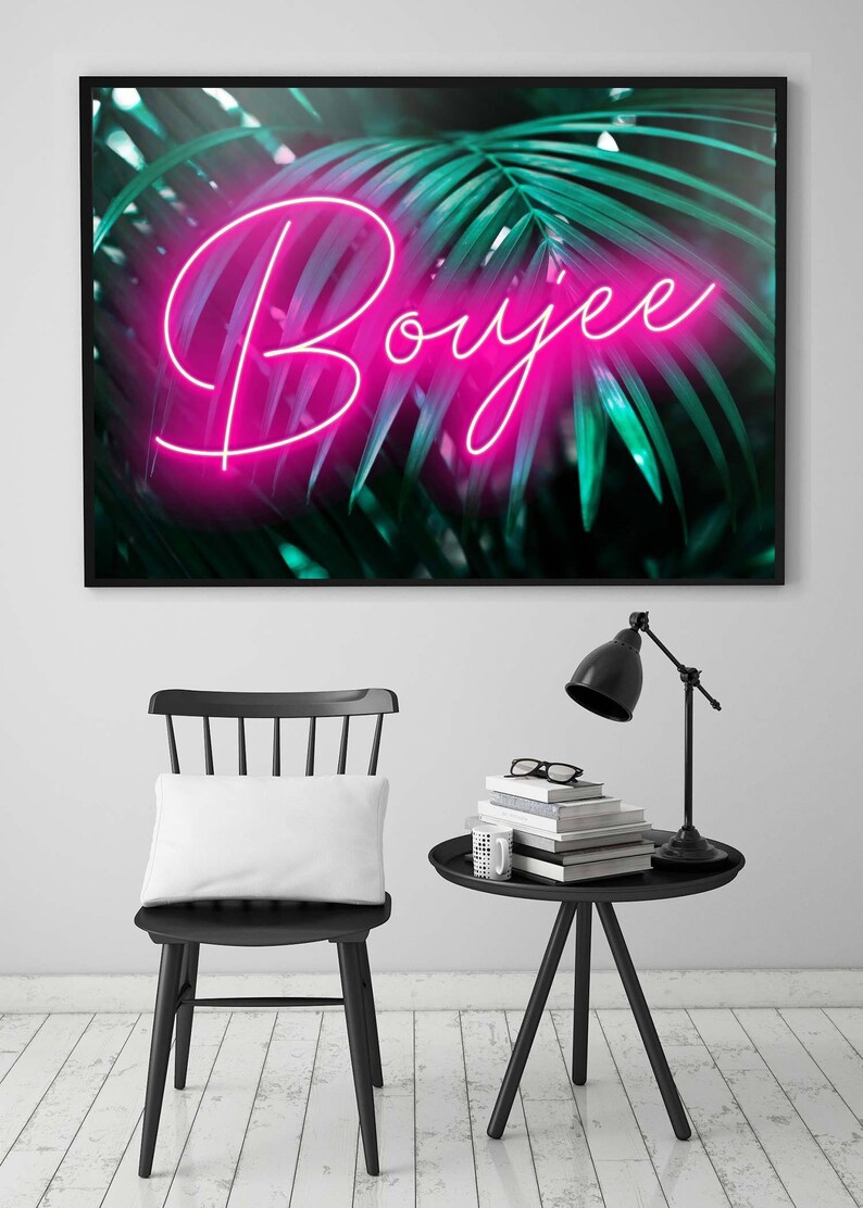 K&ouml;nnte beinhalten: Gerahmter Kunstdruck mit dem Wort "Boujee" in pinkfarbener Neon-Schrift vor einem Hintergrund aus gr&uuml;nen Palmenbl&auml;ttern. Ein schwarzer Stuhl mit wei&szlig;em Kissen und ein schwarzer Beistelltisch mit Lampe und B&uuml;chern im Vordergrund.