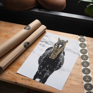 May include: A black horse with a gold-painted skull stands in a snowy field. The horse is facing the camera and has its head tilted slightly to the side. The snow is falling around the horse, and the background is a blur of white and gray.