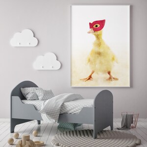 May include: A yellow duckling wearing a red superhero mask, watercolour painting on a white background, framed and hanging on a wall above a grey toddler bed with white polka dot bedding.