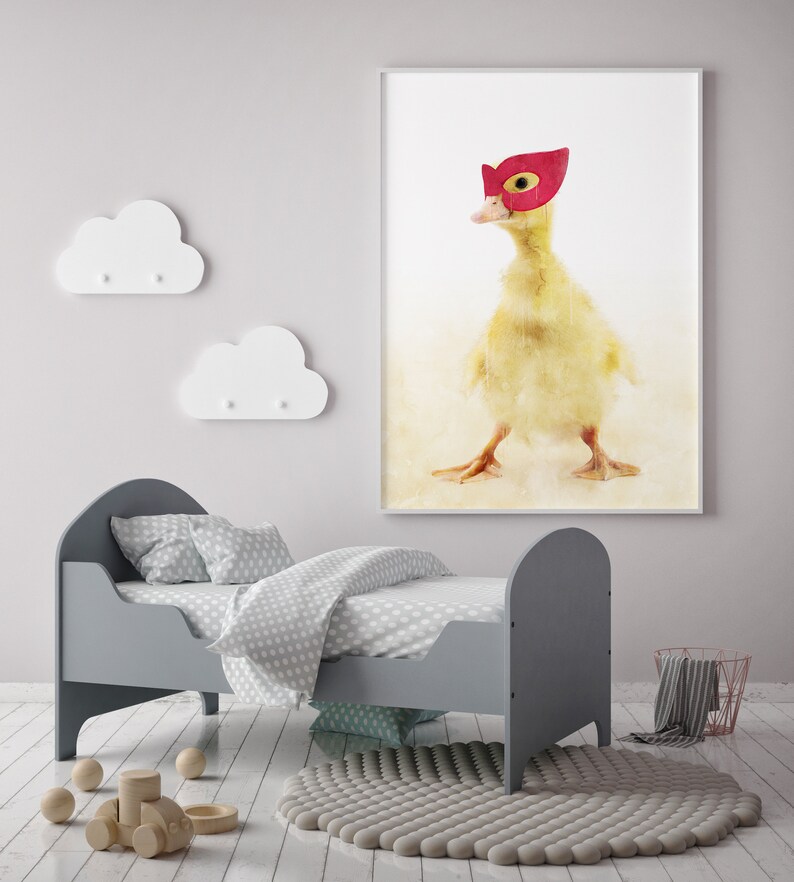 May include: A yellow duckling wearing a red superhero mask, watercolour painting on a white background, framed and hanging on a wall above a grey toddler bed with white polka dot bedding.