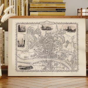 Könnte beinhalten: Gerahmte Vintage-Karte von Newcastle upon Tyne, England, in Schwarzweiß. Die Karte zeigt detaillierte Straßenlayouts, einen Fluss und Illustrationen bemerkenswerter Gebäude. Der Rahmen ist hellbeige, und die Karte wird auf einer Holzoberfläche präsentiert.