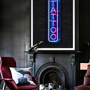 May include: A black and white photo of a neon sign that says "TATTOO" in pink letters. The sign is in front of a brick wall. The photo is framed in a white frame and is hanging above a black fireplace.
