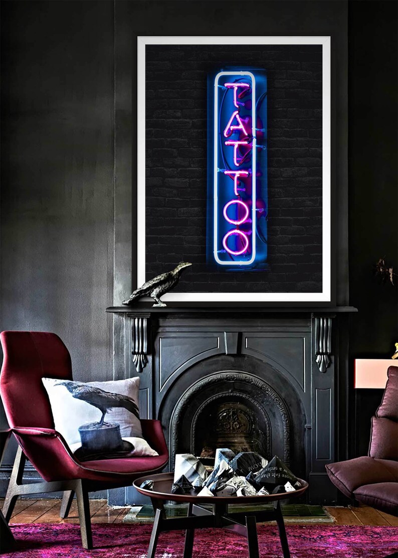 May include: A black and white photo of a neon sign that says "TATTOO" in pink letters. The sign is in front of a brick wall. The photo is framed in a white frame and is hanging above a black fireplace.