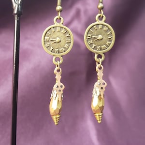 May include: A pair of antique gold-colored earrings. Each earring features a circular clock face with Roman numerals and a dangling teardrop-shaped charm. The earrings are suspended from a hook.