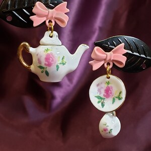 May include: A pair of earrings featuring a miniature teapot and a teacup and saucer. The teapot and teacup are white with pink floral designs. The earrings are attached to black leaves with pink bows.
