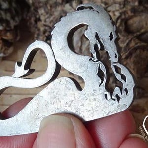 Könnte beinhalten: Ein silberner Drachenausschnitt aus Metall mit einem detaillierten Design wird in einer Hand gehalten. Der Drache ist zu einem Kreis zusammengerollt, mit seinem Schwanz im Maul.