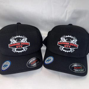 May include: Two black baseball caps with the text "HAMMER DOWN UTV" in a white and red design. The caps have a curved brim and a structured fit. Each cap has a Flexfit sticker on the brim.