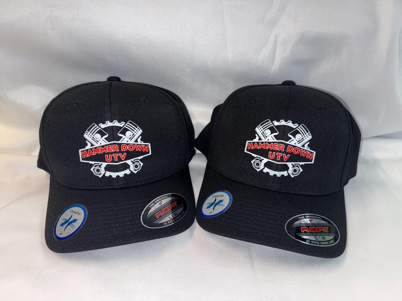 May include: Two black baseball caps with the text "HAMMER DOWN UTV" in a white and red design. The caps have a curved brim and a structured fit. Each cap has a Flexfit sticker on the brim.