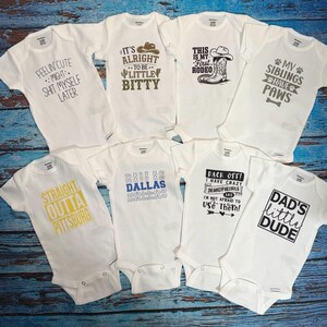 May include: A collection of white baby onesies with various printed phrases. The onesies feature text such as "FEELIN' CUTE," "IT'S ALRIGHT TO BE LITTLE BITTY," and "DAD'S LITTLE DUDE." The onesies are arranged on a blue wooden surface.