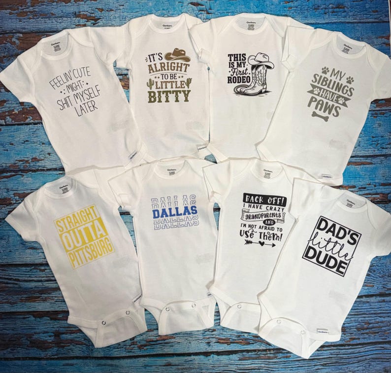May include: A collection of white baby onesies with various printed phrases. The onesies feature text such as "FEELIN' CUTE," "IT'S ALRIGHT TO BE LITTLE BITTY," and "DAD'S LITTLE DUDE." The onesies are arranged on a blue wooden surface.