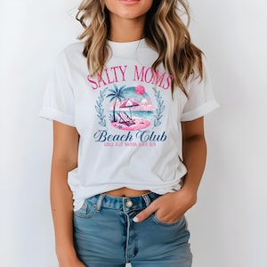 Op de afbeelding: Wit T-shirt met een roze en blauwe grafische print met de tekst "Salty Moms Beach Club Girls Just Wanna Have Sun".
