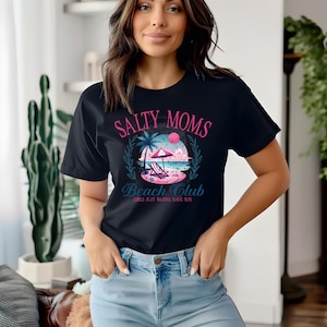 Op de afbeelding: Een zwart T-shirt met een roze en blauw grafisch ontwerp met de tekst "Salty Moms Beach Club Girls Just Wanna Have Sun".