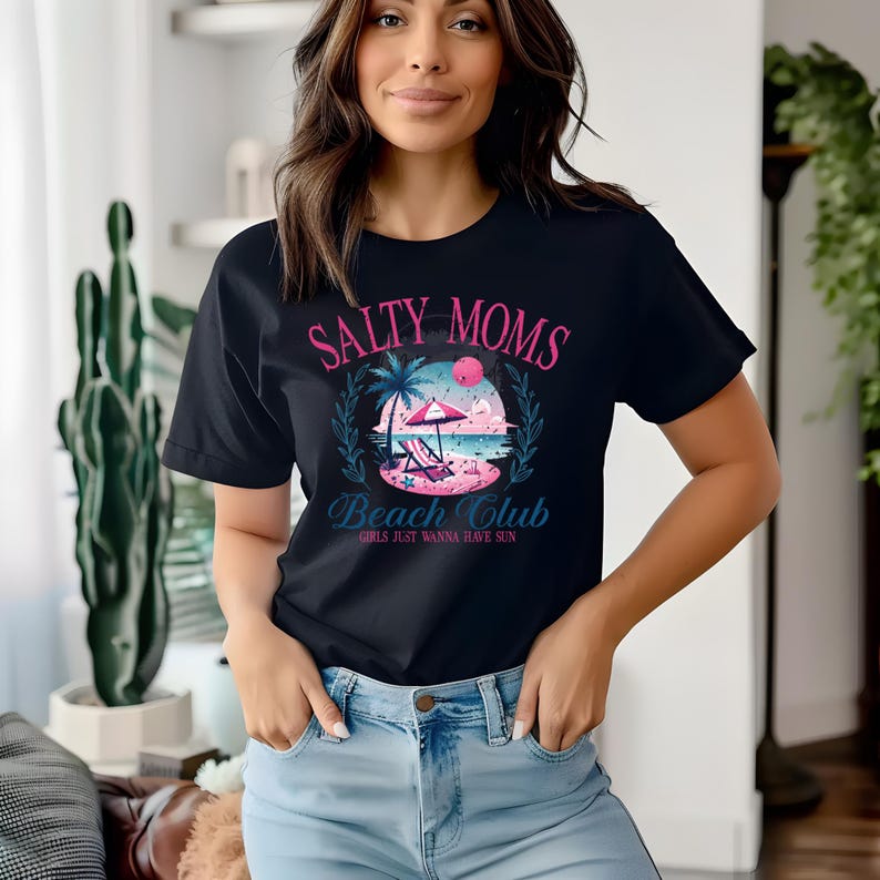 Op de afbeelding: Een zwart T-shirt met een roze en blauw grafisch ontwerp met de tekst "Salty Moms Beach Club Girls Just Wanna Have Sun".