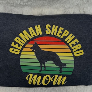 May include: Dark gray sweatshirt featuring a silhouette of a German Shepherd dog inside a colorful sunset design. The text "GERMAN SHEPHERD MOM" is arched above and below the graphic in yellow.