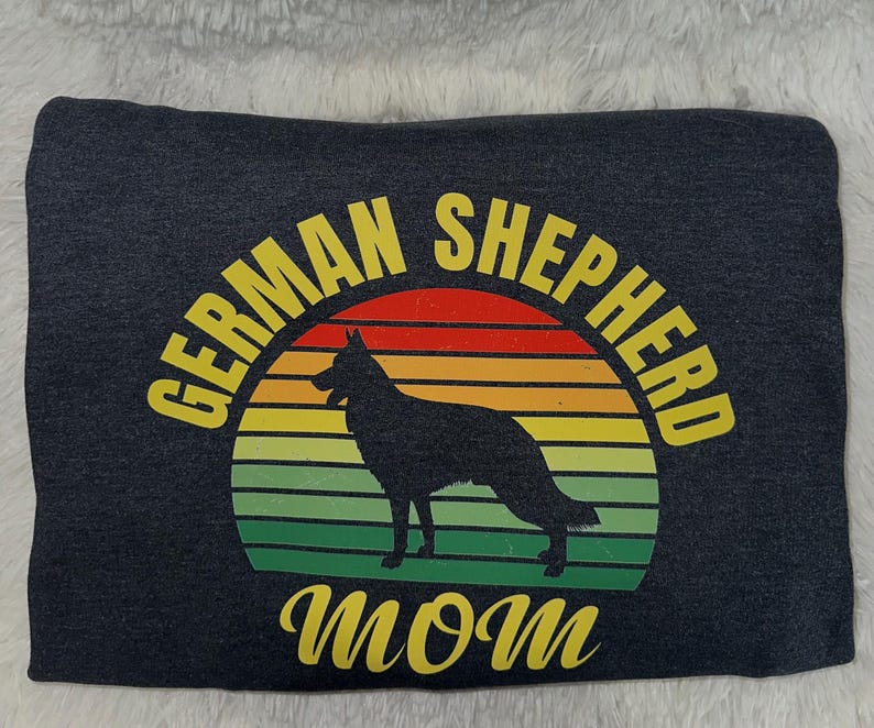 May include: Dark gray sweatshirt featuring a silhouette of a German Shepherd dog inside a colorful sunset design. The text "GERMAN SHEPHERD MOM" is arched above and below the graphic in yellow.