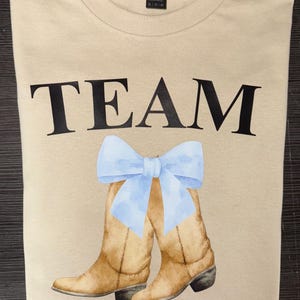 May include: Tan t-shirt with the words "TEAM BLUE" in black block letters. A watercolor illustration of brown cowboy boots with a light blue bow is between the words. The shirt is folded.