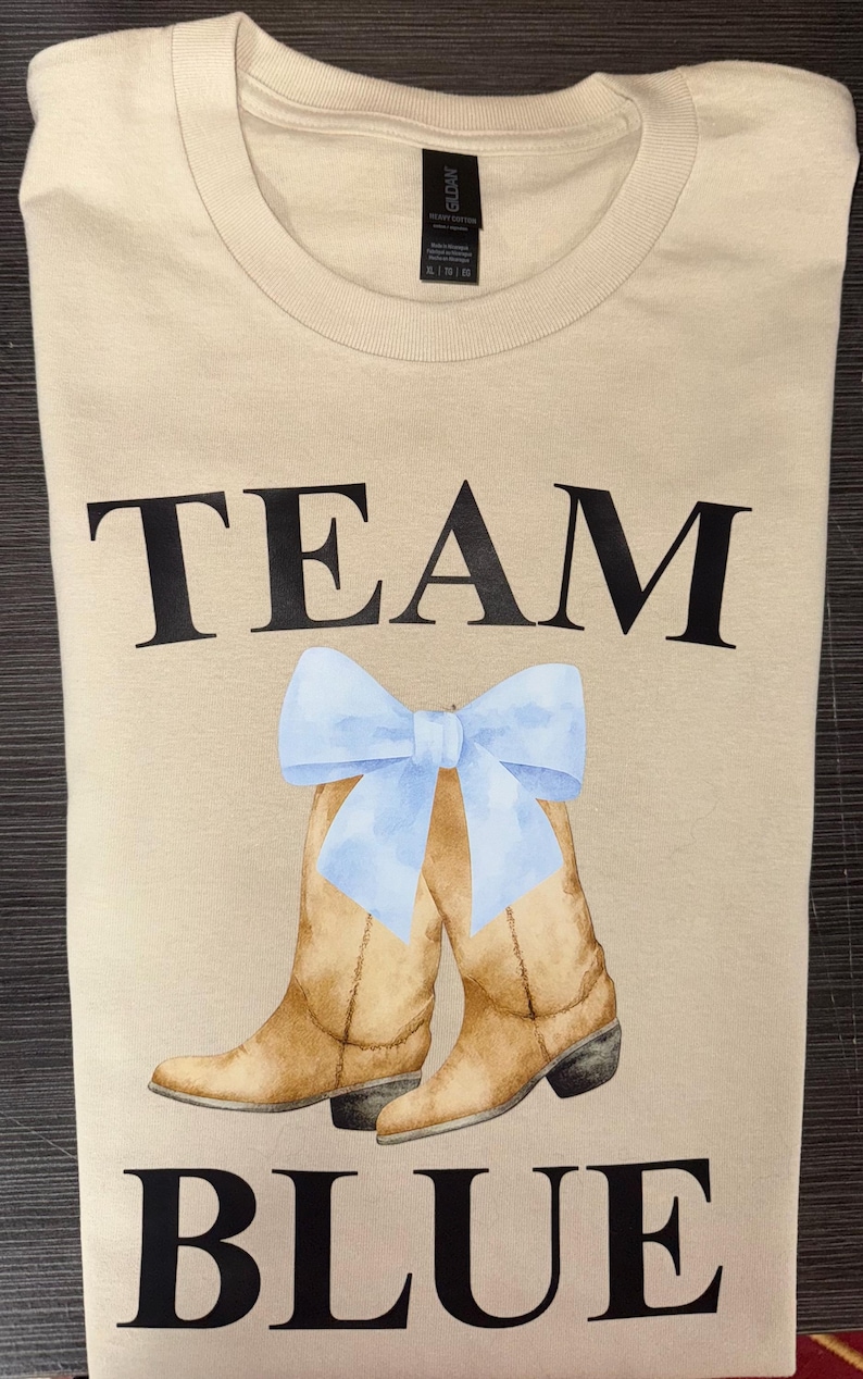 May include: Tan t-shirt with the words "TEAM BLUE" in black block letters. A watercolor illustration of brown cowboy boots with a light blue bow is between the words. The shirt is folded.