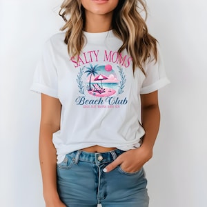 Op de afbeelding: Wit T-shirt met een roze en blauw grafisch ontwerp met de tekst "Salty Moms Beach Club Girls Just Wanna Have Sun".