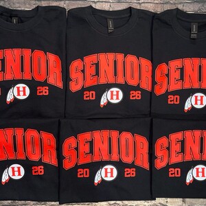 May include: Black t-shirts with the word "SENIOR" in large red block letters. Below the word "SENIOR" is the number "20" and "26" on either side of a white circle with a red "H" and a feather graphic. The shirts are likely for a graduating class.