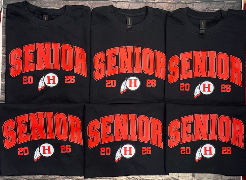 May include: Black t-shirts with the word "SENIOR" in large red block letters. Below the word "SENIOR" is the number "20" and "26" on either side of a white circle with a red "H" and a feather graphic. The shirts are likely for a graduating class.