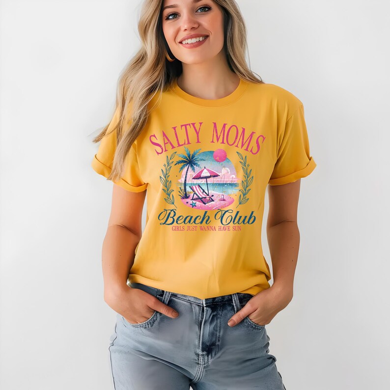 Op de afbeelding: Een geel T-shirt met de tekst "Salty Moms Beach Club Girls Just Wanna Have Sun" en een afbeelding van een strandsc&egrave;ne met een palmboom, een strandstoel en een parasol.