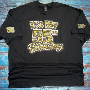 May include: Black t-shirt with the text "It's My 55th Birthday" in a yellow and black camouflage design. The number 55 is also on the left sleeve. A small logo is on the right sleeve.