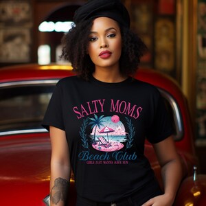 Op de afbeelding: Zwart T-shirt met een roze en blauwe grafische print met de tekst "Salty Moms Beach Club Girls Just Wanna Have Sun".