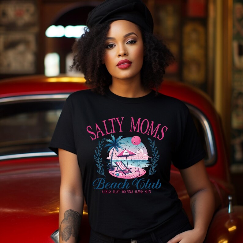 Op de afbeelding: Zwart T-shirt met een roze en blauwe grafische print met de tekst "Salty Moms Beach Club Girls Just Wanna Have Sun".