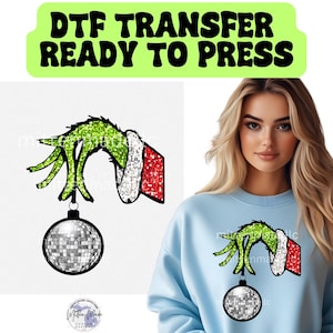 May include: DTF TRANSFER READY TO PRESS text above a green glittery Grinch hand holding a disco ball ornament. The Grinch's hand is wearing a red and white Santa-style cuff. The image is on a white background.