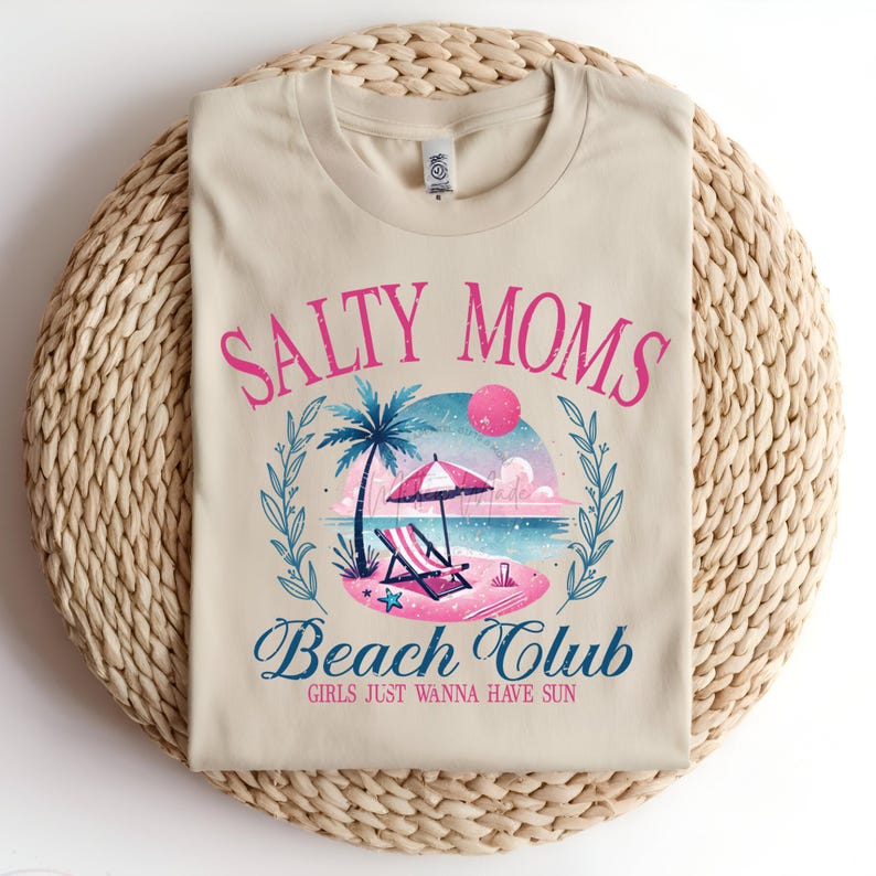 Op de afbeelding: Een beige T-shirt met een grafisch ontwerp van een strandsc&egrave;ne met een palmboom, een strandstoel en een parasol. De tekst "Salty Moms Beach Club" is in roze en blauw geschreven met de tagline "Girls Just Wanna Have Sun" eronder.