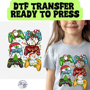 May include: A light gray t-shirt featuring a graphic print of six video game controllers decorated with Christmas themes including a Santa hat, a snowman, a candy cane, reindeer antlers, a pine tree, and colorful holiday string lights.