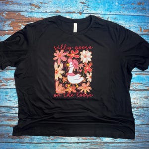 May include: Black t-shirt with a floral graphic featuring a goose wearing a pink hat and the text "silly goose on the loose" in pink. The flowers are pink, white, and orange. The shirt is made of a soft material.