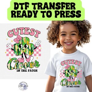 May include: A white t-shirt with a pink, green, and yellow graphic of a cartoon clover with the text "Cutest Clover in the Patch".