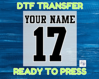 May include: A white and blue DTF transfer with the text "YOUR NAME" and the number "17" in black. The text "DTF TRANSFER" and "READY TO PRESS" are in green.