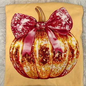 May include: A light yellow t-shirt featuring a large, sparkling pumpkin design. The pumpkin is adorned with a red bow and ribbon. The pumpkin is a mix of gold and orange sequins, creating a festive and eye-catching look.