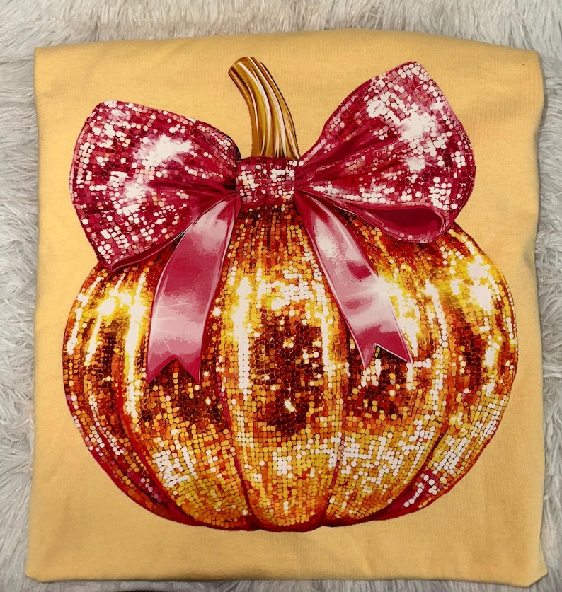May include: A light yellow t-shirt featuring a large, sparkling pumpkin design. The pumpkin is adorned with a red bow and ribbon. The pumpkin is a mix of gold and orange sequins, creating a festive and eye-catching look.