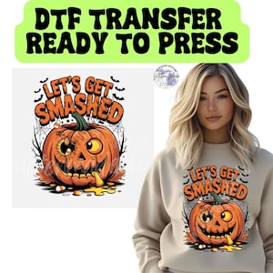 May include: A DTF transfer design featuring a cartoon pumpkin with the text "LET'S GET SMASHED" in orange and black. The pumpkin has a carved face and is surrounded by bats and spiderwebs. The design is on a light beige sweatshirt.