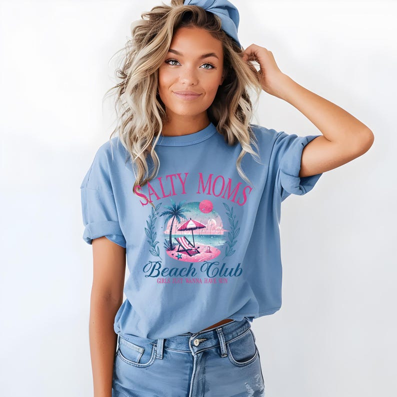 Op de afbeelding: Een lichtblauw t-shirt met een grafische print van een strandsc&egrave;ne en de tekst "Salty Moms Beach Club Girls Just Wanna Have Sun".