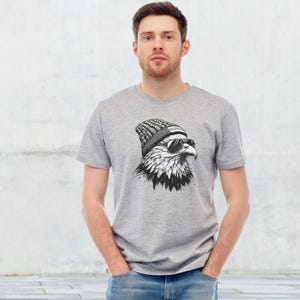 Könnte beinhalten: Ein graues T-Shirt mit einer schwarz-weißen Illustration eines Adlers, der eine Mütze und eine Sonnenbrille trägt. Die Mütze trägt den Schriftzug "FREEDOM".