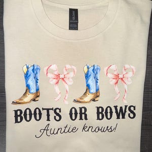 May include: Cream-colored t-shirt featuring a watercolor design of blue cowboy boots and pink bows. The text "BOOTS OR BOWS Auntie knows!" is printed below the graphic. The shirt is folded neatly.
