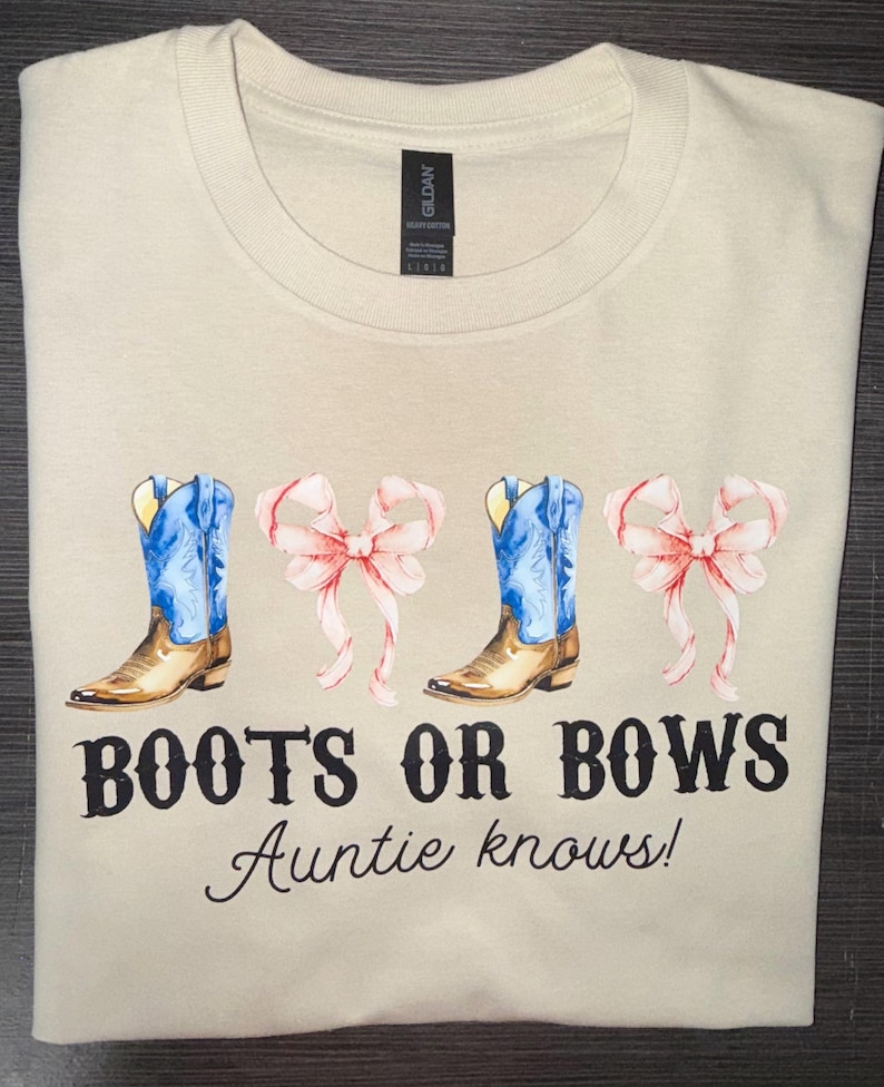 May include: Cream-colored t-shirt featuring a watercolor design of blue cowboy boots and pink bows. The text "BOOTS OR BOWS Auntie knows!" is printed below the graphic. The shirt is folded neatly.