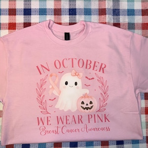 May include: Pink t-shirt with a graphic design featuring a ghost, a pink ribbon, and a jack-o'-lantern. The text reads "In October We Wear Pink Breast Cancer Awareness". The shirt is a crew neck.